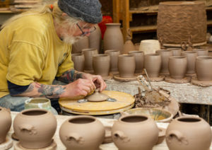 Artist in yellow shirt and grey hat shaping the lid of a ceramic pot out of clay. in the center of the imgage. In the foreground background are an array of ceramic pots