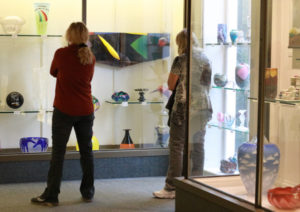 Two visitors are viewing an exhibit in the Museum of American Glass. On the right side of the image is a glass case containing glass art such as small multicolored vases and a large vase covered with pictures of clouds. To the left of this case is a case on the back wall that the visitors are looking at. There are many glass pieces of contemporary coloring and shapes.