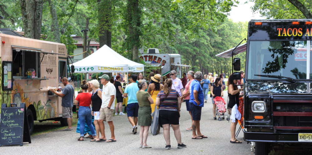 Large crowd in the center on the main street of campus. The street is lined with food trucks on the side. To the left is a vegan food truck and to the right is a tacos food trucks. There are also customers at the foodtrucks.