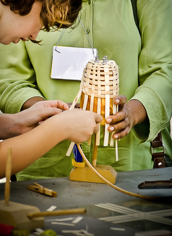 Two people one in the center behind the subject and one to the left are creating a basket in the center of the image. The subject is the base of the basket being woven on a yellow stand.