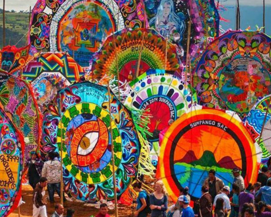 An variety of Mayan Sacred kites in different colors and sizes are presented on the grounds in the center of the image. there are about 8 kites visible in the image in three rows across the image. In the first row, on the left is a kite with a blue background and an orange eye in the center, and on the right is a circular kite with "Sumpango Sac" on a black border with mountains and an ocean painted in the center of it with a red-orange background.