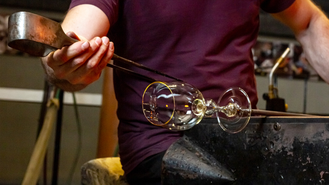 Glass artist working on a wine glass attached to a pole using tongs designed for creating glass art.