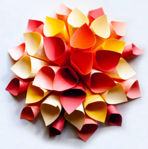 A colored varied paper sculpture made with tissue paper, cardstock, and carboard. These materials come together to create a flower shaped sculpture with a flat bottom and red, orange, and yellow petals forming the "flower"