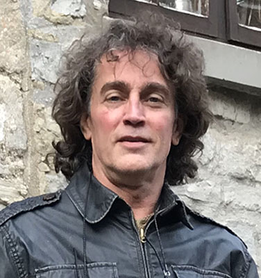 A headshot of glass artist "Gordon Smith" who is wearing a black zippered jacket and has dark curly hair and thick dark eyebrows.