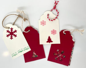 There are four Holiday Studio Sale tags in the center of the image. Two of them show the backs and two show the front. The first has three green "LET IT" At the bottom with a large red snwoflake at the top. The second back has a dark red christmas tree at the bottom and two red snowflakes above it. There are also two fronts of the tags, one with a small christmas tree at the bottom. The right front tag has a small snowflake design a t the bottom. Both designs have flannel patterns on them. Each tag has a string loop going through them. Two of the loops are light beige, one is dark brown, and one is red and white striped.