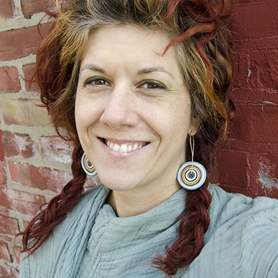 A headshot of glass artist "Amy Lemaire" who has red pigtails resting on her blue shirt and red and blue multicolored earrings that appear to look like yo-yo's. She also has brown eyes and is smiling at the camera.