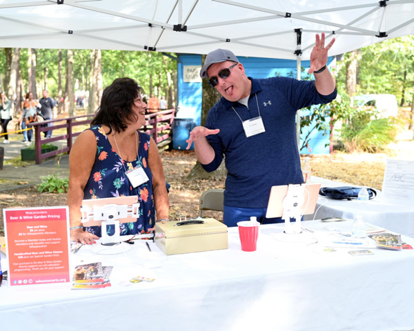 Two volunteers in the Beer and Wine Garden at the Festival of Fine Craft smiling and having fun before visitors arrive. They are standing behind a white table with a red sign on the left, two ipads set-up on either side for payment, and a box in the middle. Both volunteers are wearing nametags and the woman on the left has dark hair and is wearing a blue flowery dress. She is smiling at the man with a light blue hat, sunglass, and is smiling goofingly at her with his hands in the air. He is wearing a blue long-sleeve shirt over a white shirt and has blue jeans on. There is a blue building in the back and three people walking on the left side of the background.