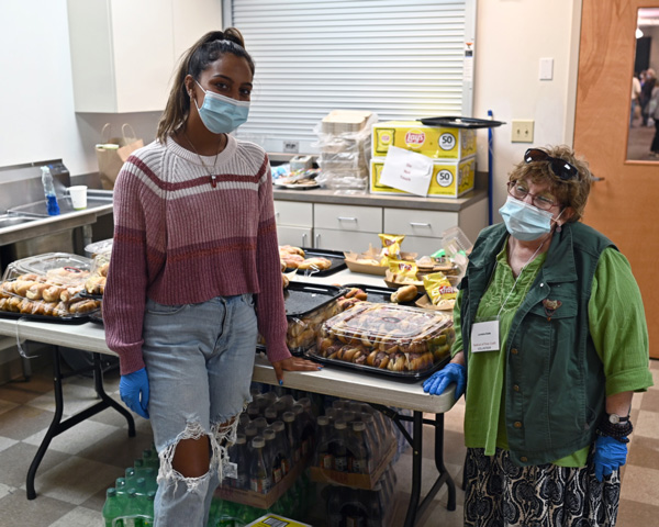 A picture of two volunteers at the Festival of Fine Craft in front of all the volunteer refreshments. Both are wearing masks and blue gloves. The young woman on the left is wearing a pink, white, and maroon layer pattern sweater. She is also wearing ripped jeans and a necklace. The older woman on the right is wearing sunglass on her head and is wearing regular glasses. She also has a nametag around her neck and is wearing a green shirt with a darker green vest over top it.