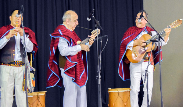 Three men with grey hair, light pants, and a red garment over their shoulders are playing instruments. The first one is playing a flute, the middle man a guitar, and the third man a guitar as well. There are two drums in the background.
