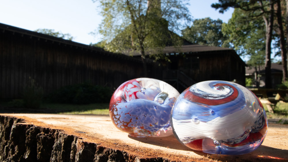 Two red white, and blue paperweights placed side-by-side on a tree stump with the WheatonArts glass studio behind it.