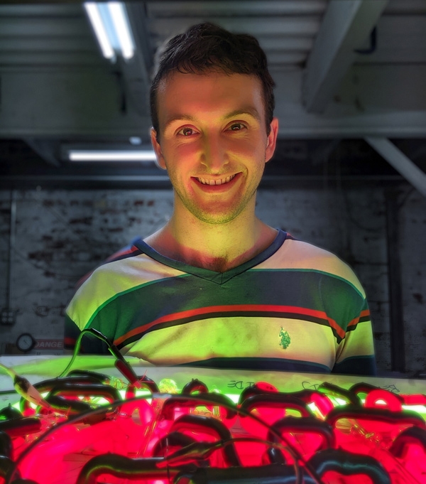 James Aker wearing a green, red, and white striped shirt smiling at the camera behind one of his neon glass sculptures.