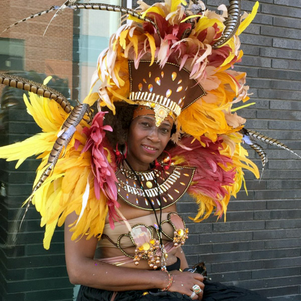 Mafalda Thomas Bouzy, a performer, is sitting on a bench and smiling at the camera. She is wearing a yellow and pink festive hat-like garment.