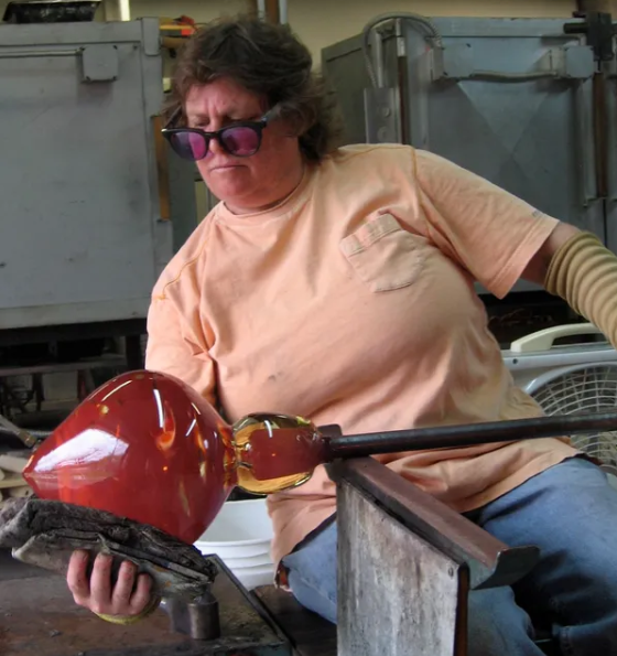 A woman wearing safety glasses, wearing a peach colored shirt, sitting on a bench, is holding a rode with a large glass piece on the end in her left hand and is shapping it with a cloth in her right hand.