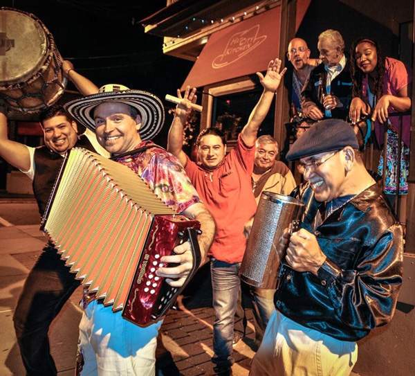 Viva Vallenato Cumbia Band performing outside ay night with a large sign in the background. The band has four men holding instruments posing for the camera. There is also a man behind them and three people watching over the railing.