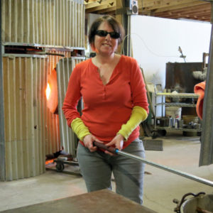 Alison Ruzsa, a middle-aged woman in a red long-sleeve shirt and safety goggles, is smiling at the camera while holding glass with a rod in a furnace. There is another furnace behind her.