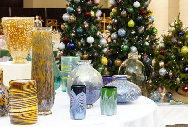 Items from the Holiday Studio Sale on a white table. There are three christmas trees with multicolored ornaments. The tree to the right is smaller than the rest. Four of the items are dark blue, three are orange, and three are green. One of the orange ones is one a raised centerpiece of the table.