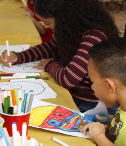 A young boy in a yellow shirt, sitting at a brown table, is cutting out his drawing of a bright-colored skull. To his right, the back of the image is a girl with long hair and a red-striped shirt, coloring in a skull template with an orange marker. There is a stack of skull template paper between them. At the bottom left corner of the image are three cups containing a variety of colored markers.