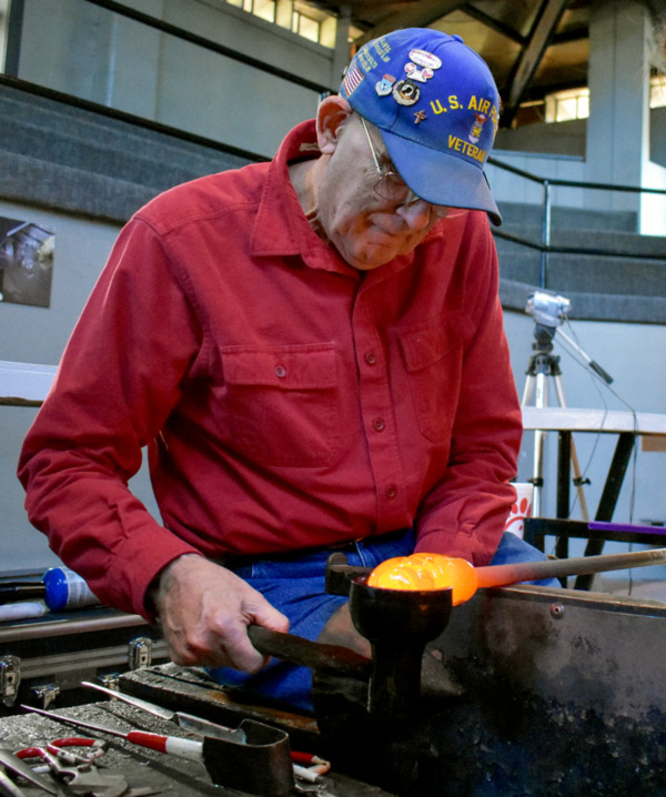 A glass blowing class for the Veterans Day Make-Your-Own Experience. He is wearing a red shirt, blue jeans, and a blue U.S. Air Force Veteran hat. He is sitting on a bench in the glass studio holding a glass blowing rod with hot glass on the end with his left hand and shaping it with a metal cup-like tool with his right.