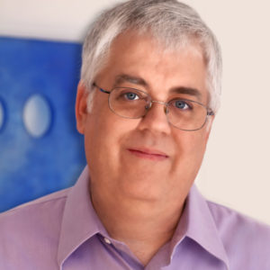 A closeup of JG Harrington from the shoulders up. Her is wearing a purrple collared shirt, glass, and has short grey hair. There is a blue square art piece behind him.