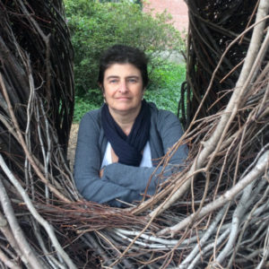 A medium close up from the arms up of Jere Gibber. Gibber is wearing a blue scarf and a grey long-sleeve shirt. The subject is surrounded by a lot of sticks creating a frame.