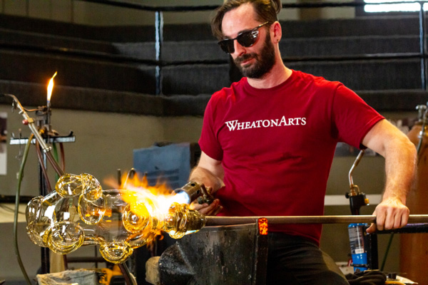 A medium close up of Alex Rosenberg, dark hair, sunglasses, and a red WheatonArts shirt, as he is holding a blow rod with a large yellow piece attached to it. In his right hand he is heating up the glass with a blowtorch.