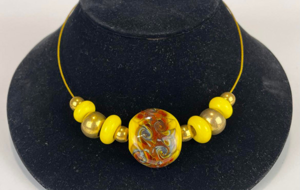 An extreme close-up of a yellow bead necklace by WheatonArts flameworker, Chris Coffman, on a black jewelry display. The string of the necklace is yellow, there is one large yellow bead with brown and blue patterns faded onto it. To either side of the necklace are three gold beads with yellow beads between them. The outside beads on each side are smaller than the others.