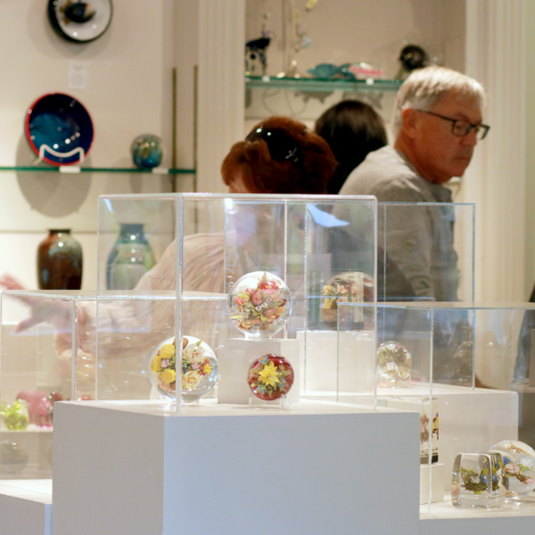 A close up of three flower paperweights in a square glass case in the gallery. Two of the paperweights are larger, clear, and have light colored flower pedals. The third paperweight, at the bottom right of the case, is small, red, and has a yellow flower in the center. There are two people in the background, one is a woman with red hair, sunglasses, and is waving her hand. Her face is not visible. To her right is a man with grey hair and dark glasses. On the wall behind them are two large glass shelves with a variety of glass and ceramic pieces.
