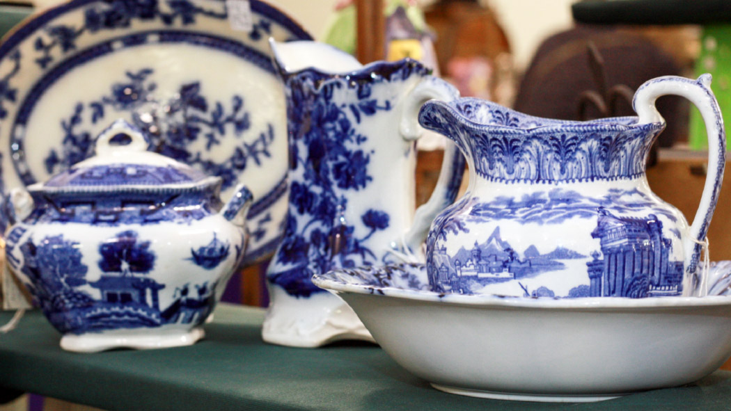 An extreme close-up of a ceramic tea set. The set consists of 4 pieces: a large plate, a tea kettle, a pitcher, and a tea pot in a bowl. All of these items have intricate blue architectural designs with white backgrounds.