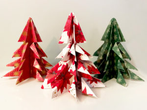 Three paper Holiday themed trees center the image. The first one is red with white dots and small yellow sun designs. The one in the middle is white with large red flowers as a pattern. It also has small red dots all over it. The tree on the right is a traditional green with a snowflake design all over it.