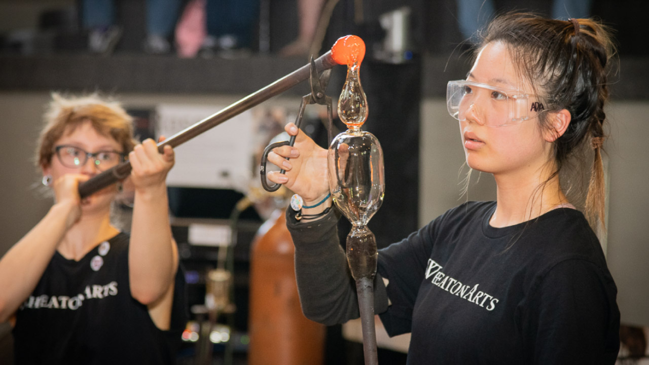 Image of WheatonArts Glass Studio interns working in the studio. Each intern is wearing glasses and a black WheatonArts shirt. The intern closest to the camera is holding a metal rod with clear glass at the top in front of their face. The other intern is holding another glass rod by their face angled so that the orange hot glass at the top is being poured on top of the clear hot glass in a tear drop shape. The intern closest to the camera is holding a pair of metal pliers to hold the rod that is pouring the glass steady.