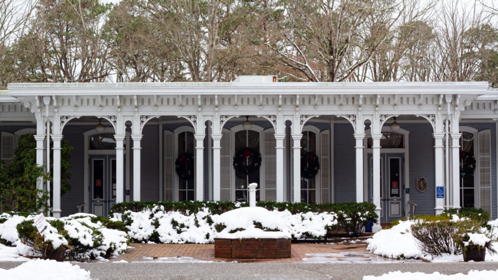 A wide shot of The Museum of American Glass coated with snow and decorated with holiday wreaths. The museum is a large pale blue. The sign in the middle of the image is seen from the side and is covered in snow. In front of the museum are bushes that are also covered in snow.