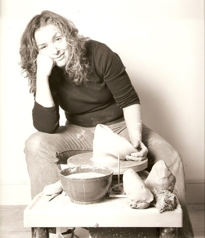 A vertical medium close-up of Jackie Sandro Greenwell, wearing a black long-sleeve shirt and clay-covered jeans, is spinning clay. The image is in black & white clay, and this image is a headshot for her Wheaton Conversations virtual session.