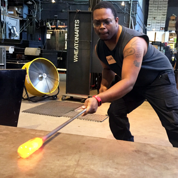 A workshop student with tattoos on their left forearm, rolling hot glass on a flat metal surface in the WheatonArts' glass studio.