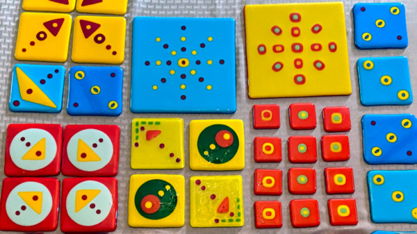 An image of several square fused glass panels. Six yellow & medium panels with arrows and orbs & one large panel with several small orange & blue dots are throughout the display. Four medium red panels with yellow triangles & red circles forming an arrow, are at the bottom left corner, twelve small red panels with yellow rings, green & yellow orbs, and yellow & green orbs are at the bottom right corner. Six medium blue panels, with yellow & blue dots, are on both sides of the image. A large blue panel with yellow & blue orbs is at top of the image.