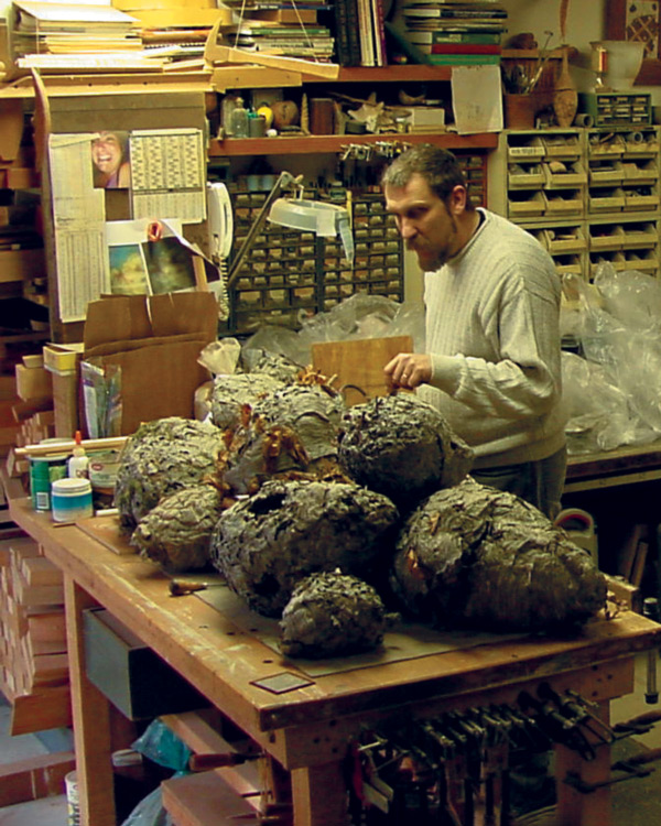 A vertical image of Jim Greenwell, a tall brown-haired man wearing a white sweater, is holding his hand above several large grey pieces for a sculpture. He is standing to the right of the table in the center of the image.