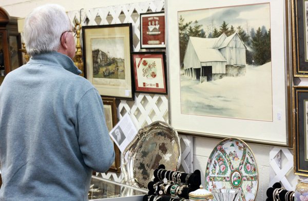 A midde-aged man with silver hair and glasses, wearing a light blue sweatshirt, is standing with his back to the camera in front of a collection of antique paintings and plates. At the bottom of the image are two large multicolored plates, and the one on the right has pink & blue flowery orbs. At the top of the image is a large painting of a shed surrounded by snows and trees, next to a variety of smaller paintings.