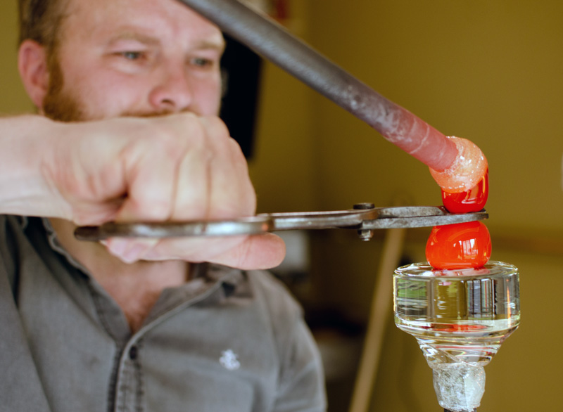 A close-up of a WheatonArts Paperweight fest breakout sessions artist William Manson, using a tool to attach two small hot glass globs to a clear glass piece.