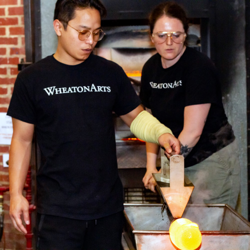 An image used for the WheatonArts glass studio internship program is of two WheatonArts glass studio artists creating a glass piece by rolling it on a surface. They are both wearing WheatonArts shirts.