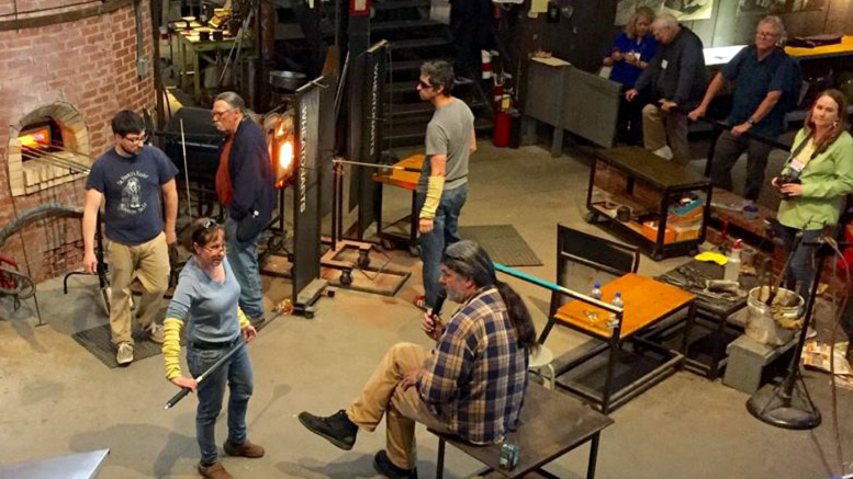 Paperweight fest demonstrating artist, Alison Ruzsa, is standing in the center of the WheatonArts' glass studio. Several people are assisting in the studio, a man is on the bench with a microphone, and several people are in the audience.