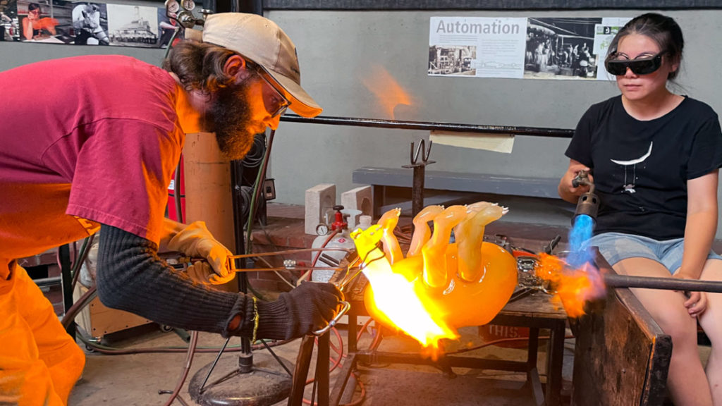 Visiting artists are renting out of the glass studio space and & equipment. Two people are working on a glass piece with torches at a workbench in the glass studio. The piece appears to be a pig.