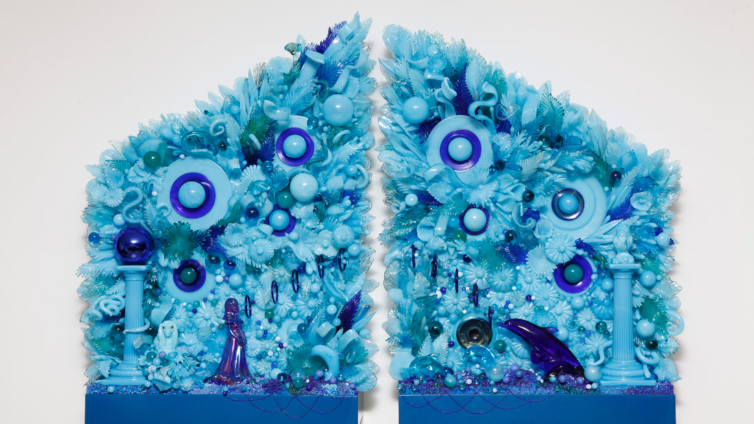 A sculpture by the Alchemy of Adornment exhibit artist Amber Cowen is in two blue symmetrical parts. The backgrounds of each glass sculpture are flowery and have several eye-like figures. At the bottom of the left section is a figure of a woman, and on the right, a bird.