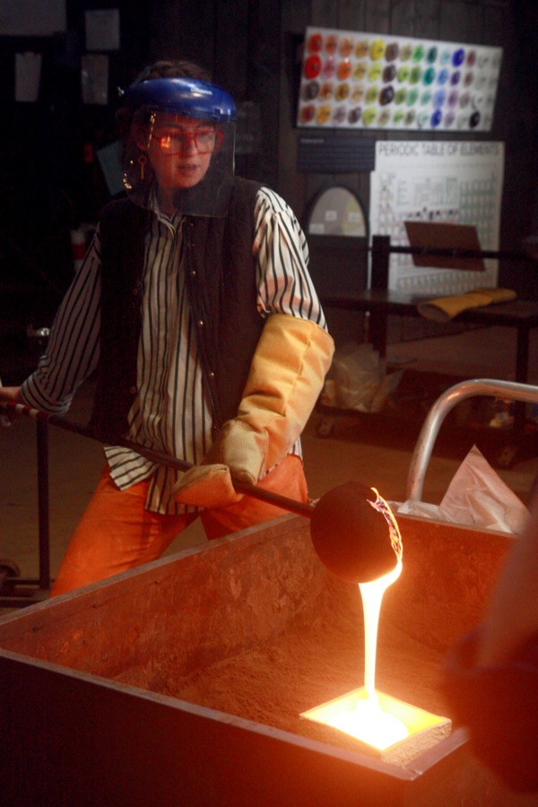 Grace Whiteside, a glass worker, is renting out the glass studio space & equipment. They are wearing a safety helmet while holding a large spoon-like tool used to manipulate hot glass. There is bright hot glass pouring out of the tool.