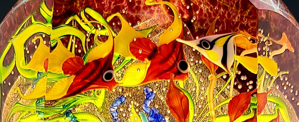 A close-up of a bright multi-colored paperweight with ocean images contains two orange-striped fish and a white & black striped fish. There is multi-colored plant life between the fish.