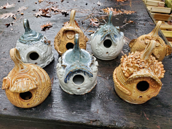 Seven ceramic birdhouses/birdfeeders by ceramic workshop instructor Phyllis Seidner are presented on a dark table and have two holes on each item's front side. Three are blue & and four are yellow.