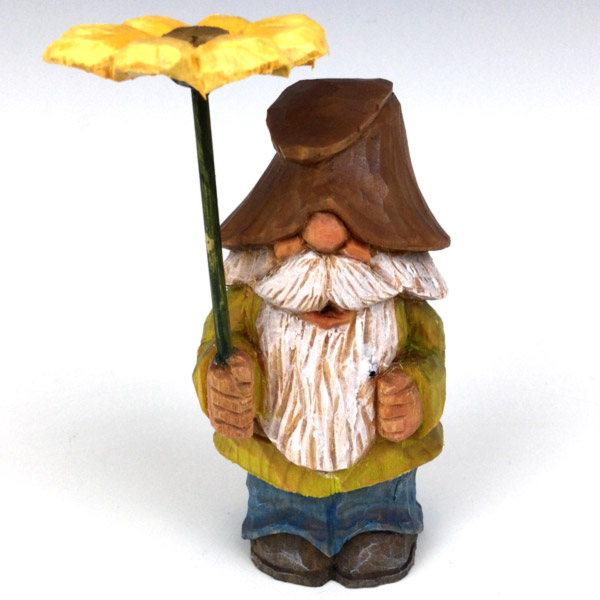 A wood-carved gnome by local artist Domenick Maggio is displayed over a white background. It is holding a yellow flower, "wearing" a green painted shirt, and has a long white beard, with a floppy brown hat covering its eyes.