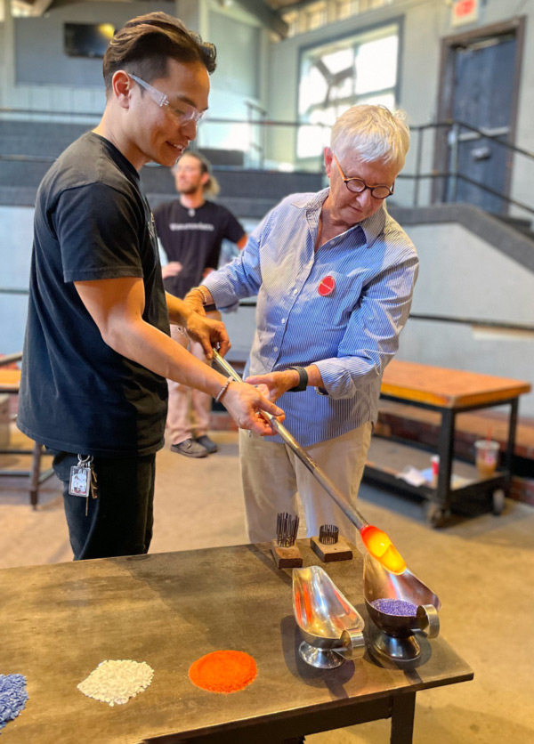 A glass studio make-your-own experience instructor, on the left in a black shirt, is assisting a man in a blue shirt, on the right, in covering the glass on the end of his rod with color.