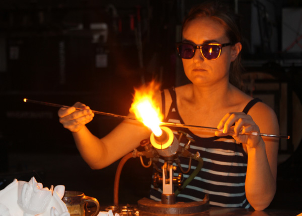 Alchemy of Adornment exhibit artist Amber Cowen, wearing safety goggles, is fusing glass on two small rods in front of a flame. She is wearing safety goggles and is to the left of the torch.