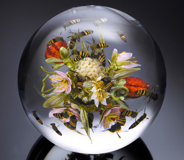 This detailed paperweight by Paul Stankard contains a scene of several glass bees flying around a variety or bright-colored flowers.