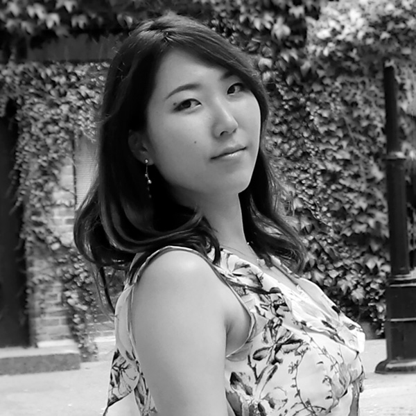 This image is a grey-scale headshot of 2023 creative glass fellow Eriko Kobayashi, who is wearing a flower print dress, has wavy dark hair, and is smiling at the camera.