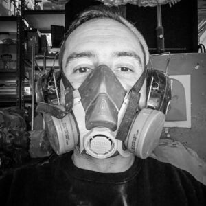This image is a headshot of 2023 creative glass fellow Esteban Salazar, who is wearing a breathing mask used for working with chemicals and a black shirt.
